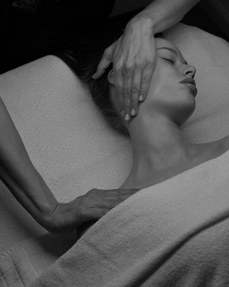 A woman receiving a head massage with her eyes closed, lying on a treatment table covered with a towel, with hands gently massaging her face and neck.