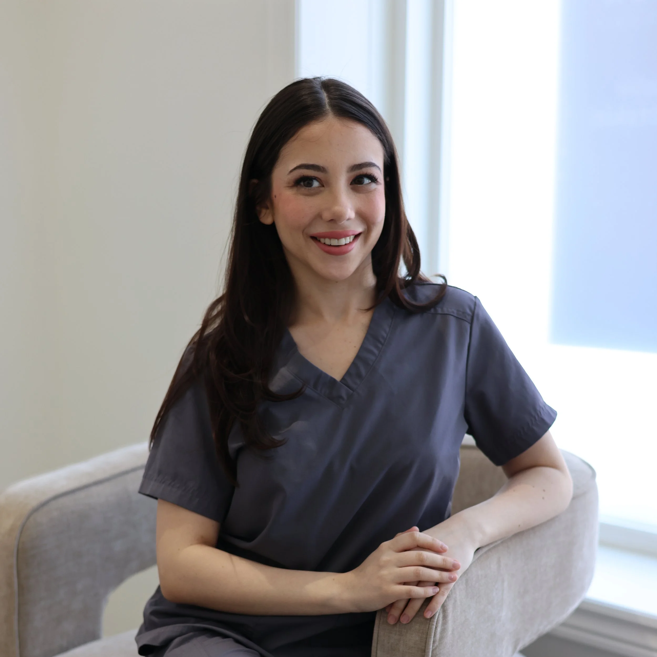 A young woman with long dark hair and light skin, wearing scrubs, smiling and leaning on a beige couch with her arm crossed, in a bright room with natural light.