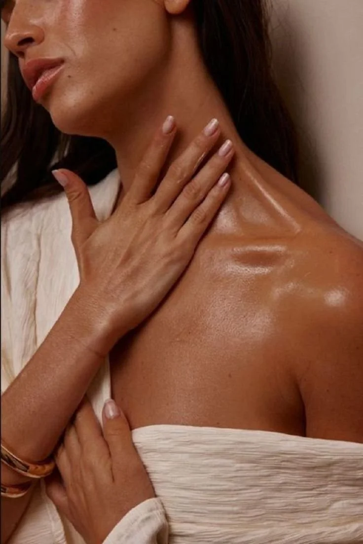 Close-up of a woman's shoulder, neck, and hand, with the woman touching her neck and chest, featuring smooth, glowing skin.