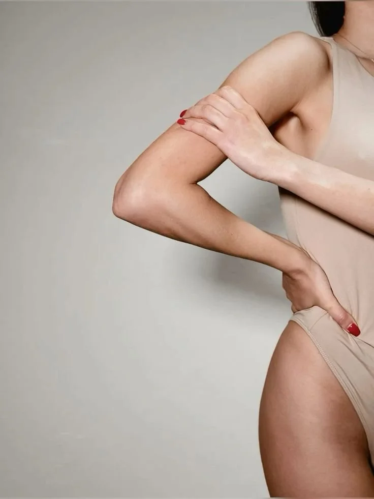 A woman wearing a beige sleeveless top, with her right arm crossed over her chest and her left hand on her hip, posing against a plain background.