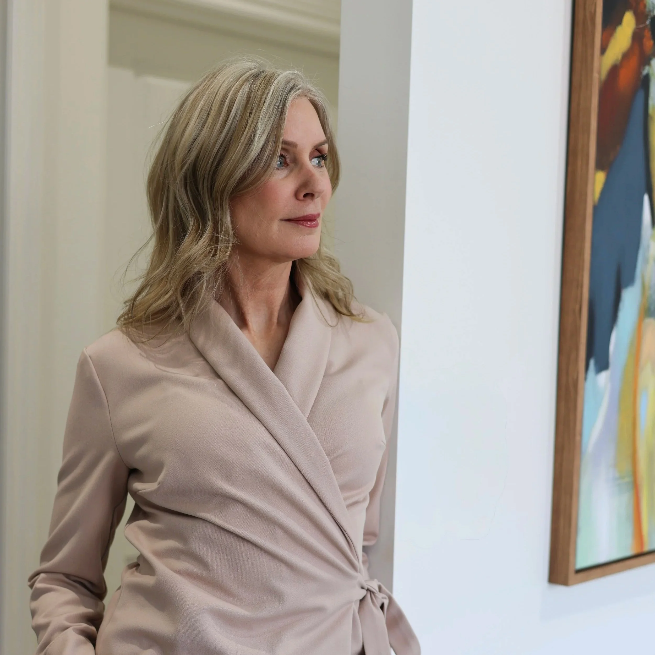 Close-up of a middle-aged woman with blonde hair and blue eyes, smiling, standing in front of colorful abstract artwork on a white wall.