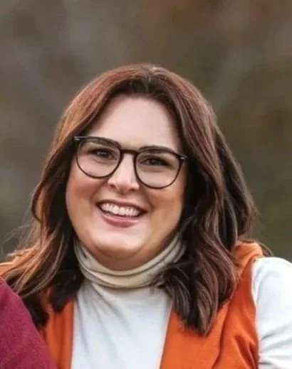 A woman with brown hair and glasses smiling outdoors, wearing a white turtleneck and an orange vest.