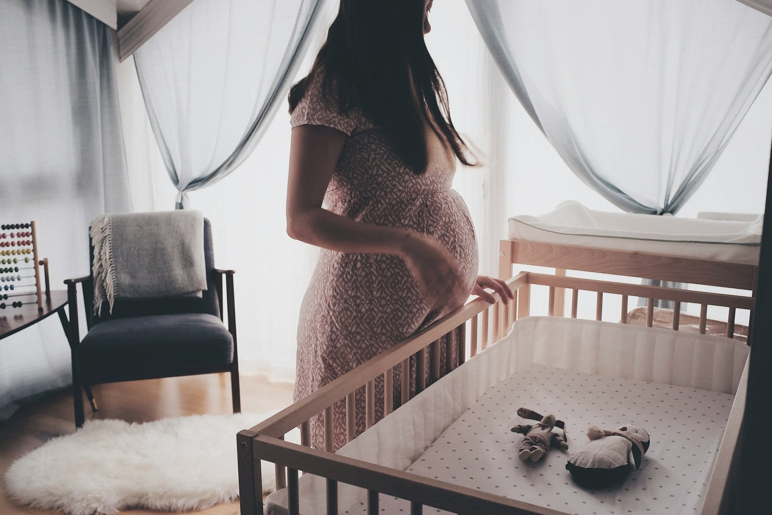A pregnant woman standing beside a crib in a cozy, well-lit nursery with sheer curtains and a plush white rug.