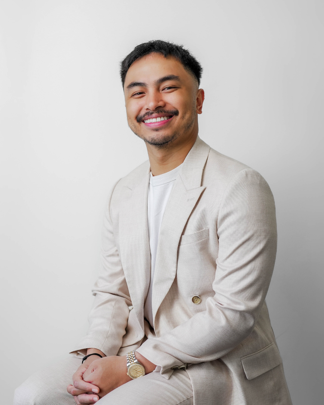 A smiling man with short black hair, a mustache, and beard, wearing a light beige suit, white shirt, and a gold watch, sitting against a plain white background.