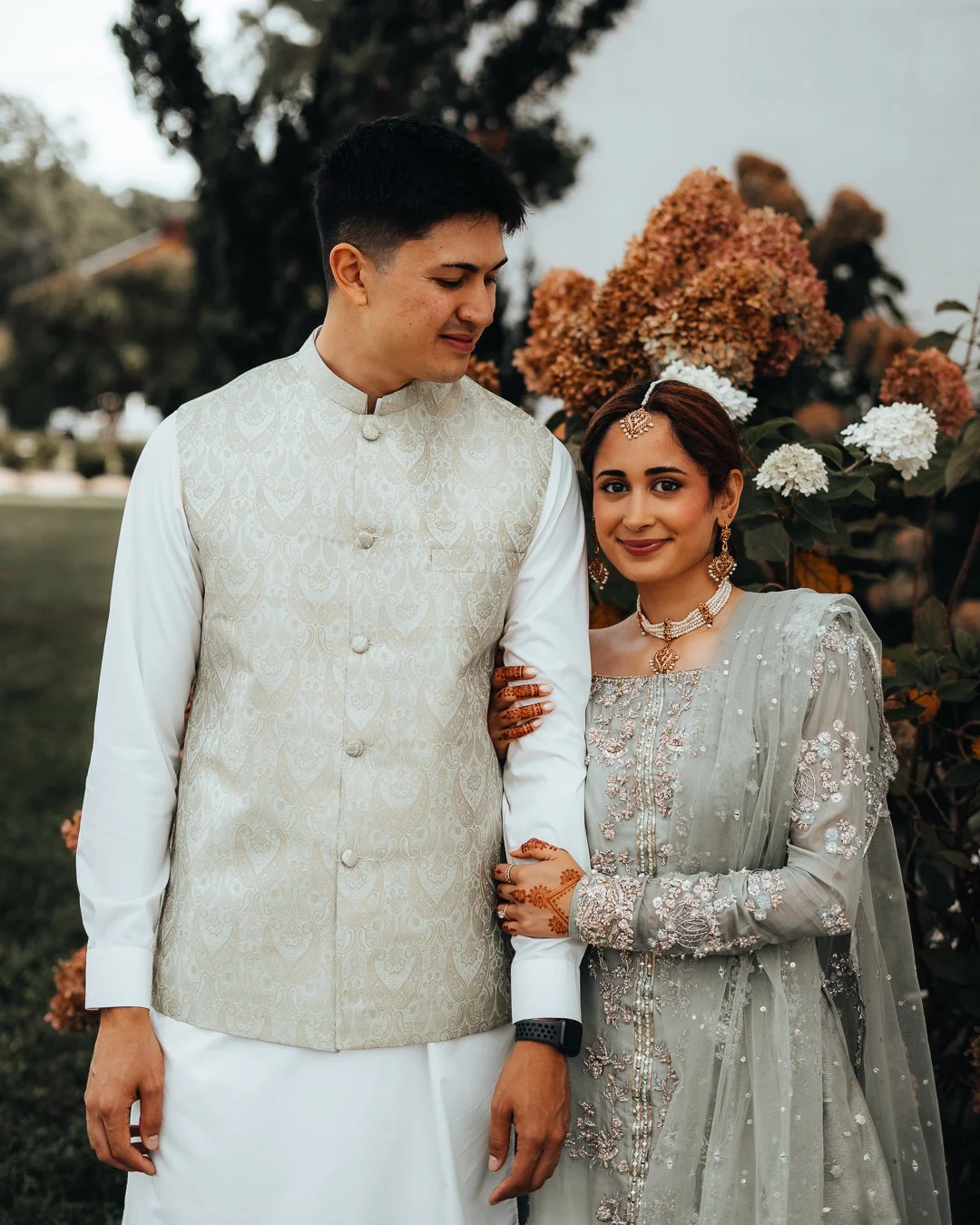 A couple in traditional Indian attire standing outdoors with flowers in the background, the woman is smiling and the man is looking down.