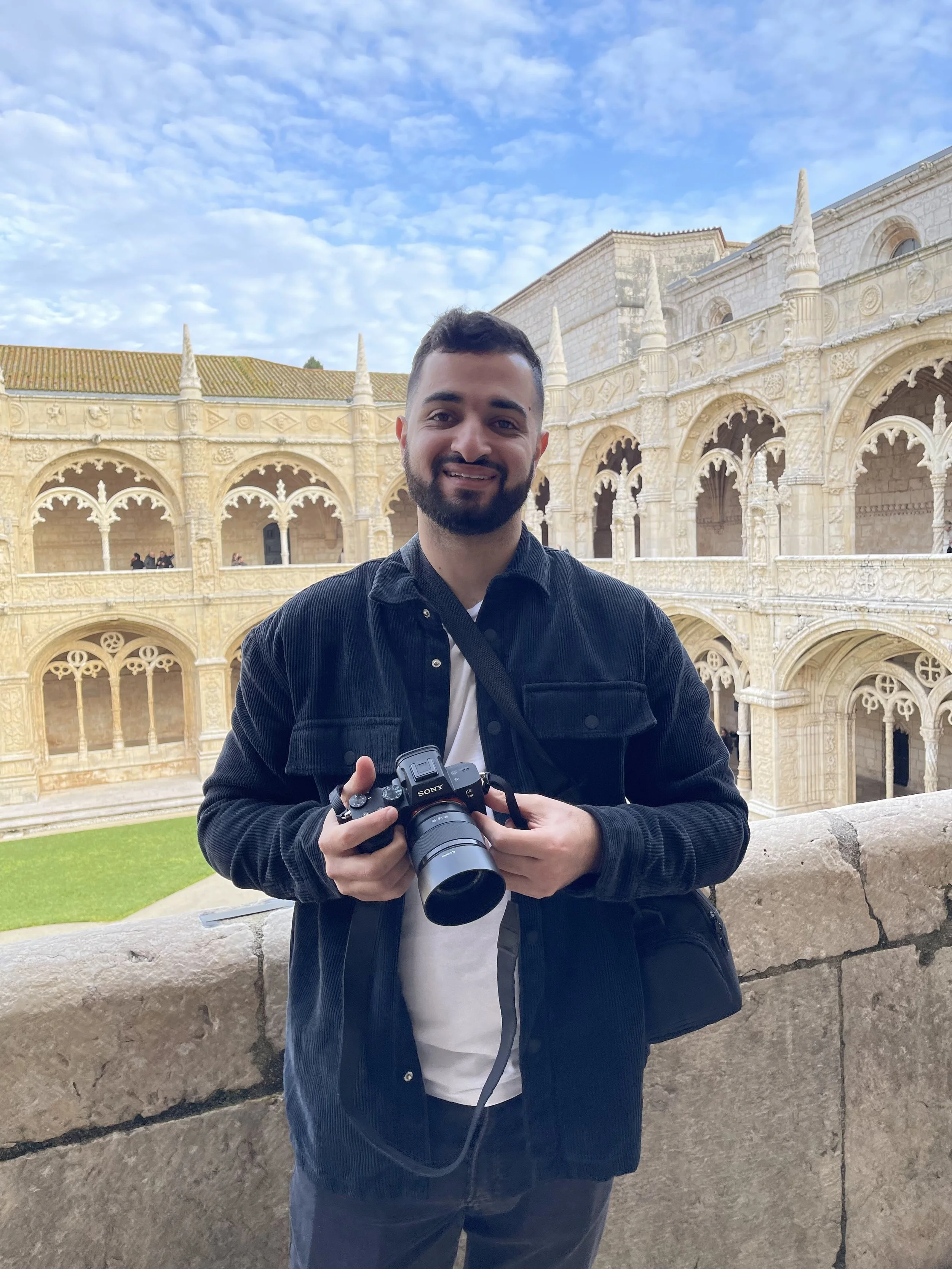 A man with a beard and short dark hair holding a camera, standing outdoors with an ornate, historic stone building in the background, under a partly cloudy sky.