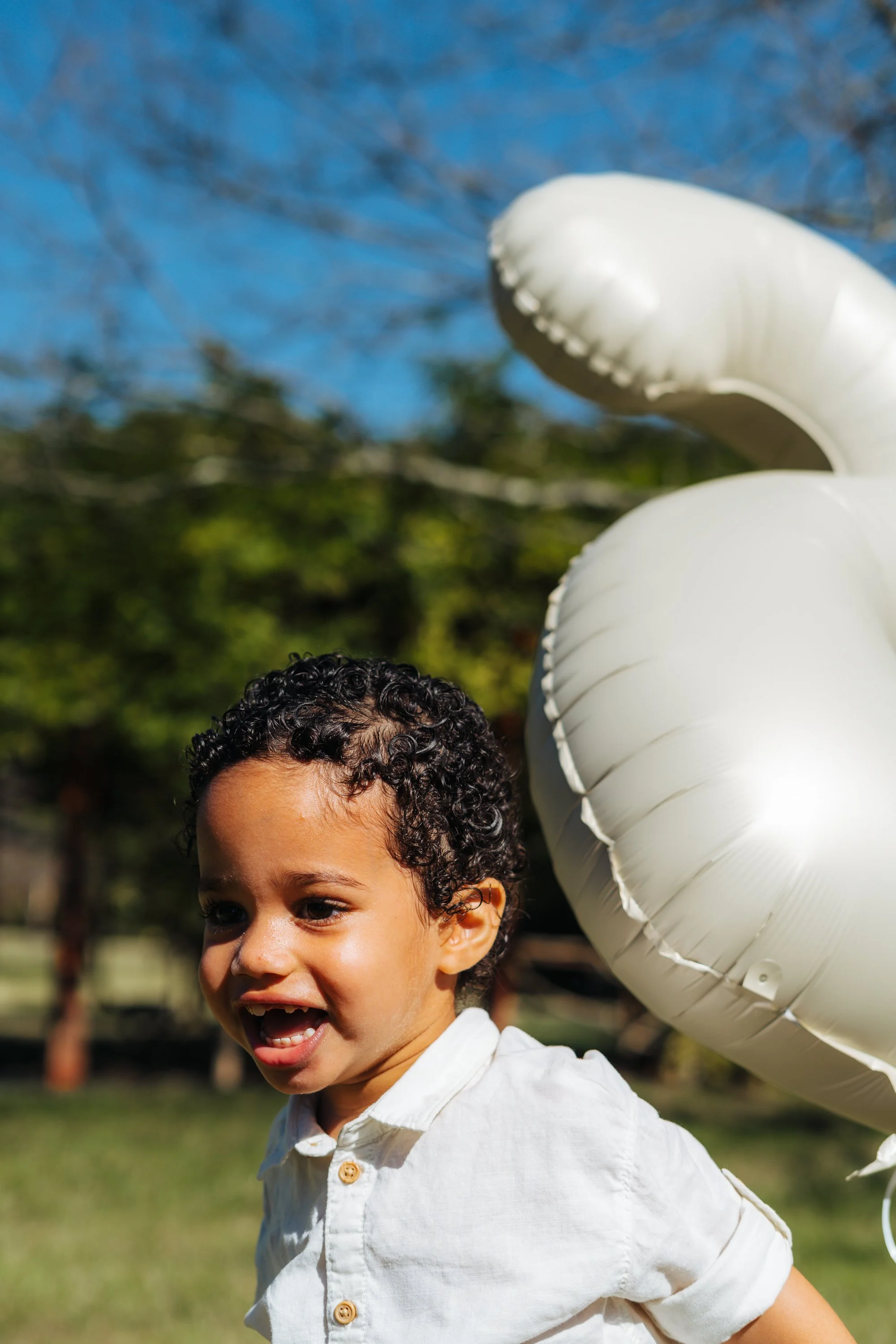 Young boy with curly hair smiling outdoors next to a white swan-shaped balloon.