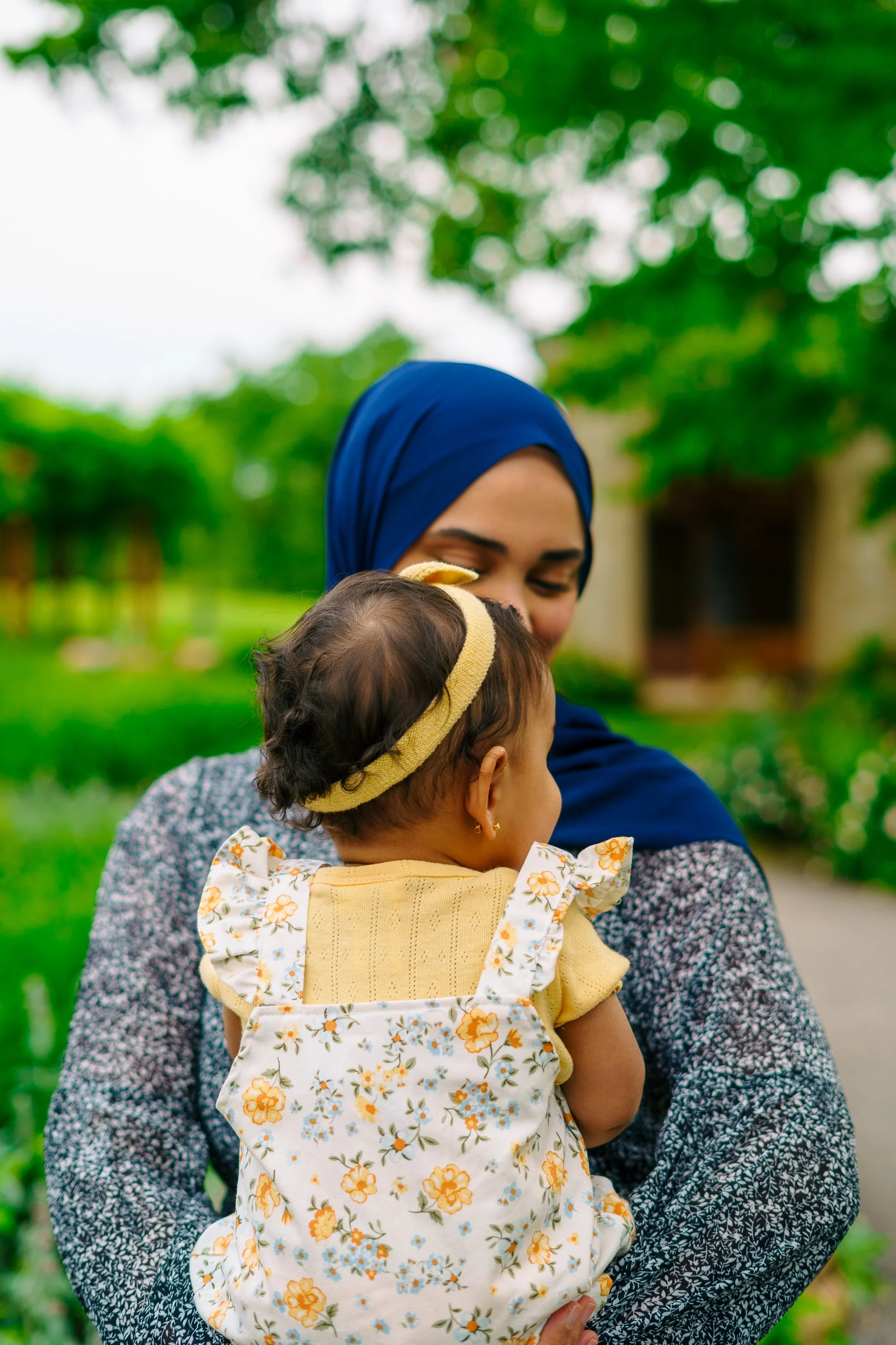 Woman wearing a blue headscarf holding a baby in a floral carrier outdoors with green trees and a house in the background.