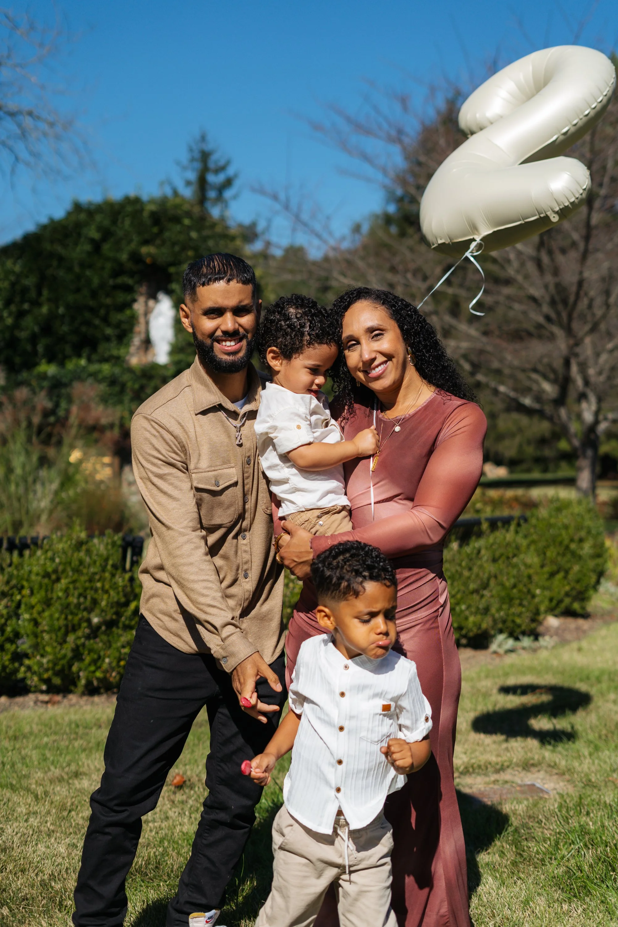 A family of four celebrating outdoors with balloons, with a woman holding a curly-haired child and a man holding hands with a boy, all smiling, in a grassy park with green bushes and trees in the background.