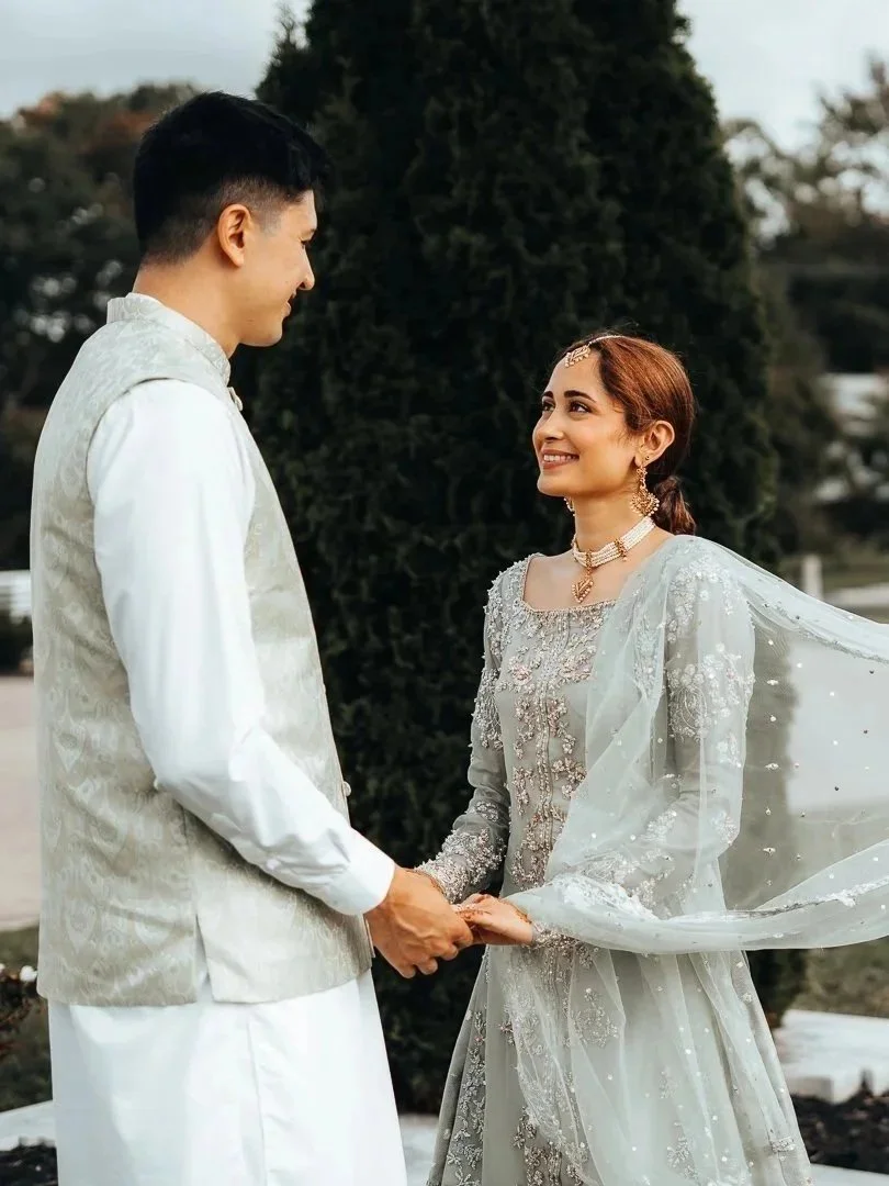 A bride and groom holding hands outdoors, dressed in traditional wedding attire, smiling at each other.