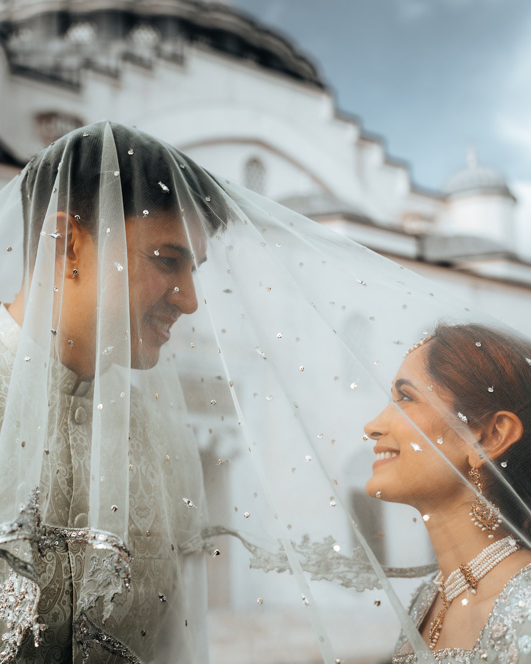 A bride and groom face each other under a veil at their wedding, with a white building in the background.