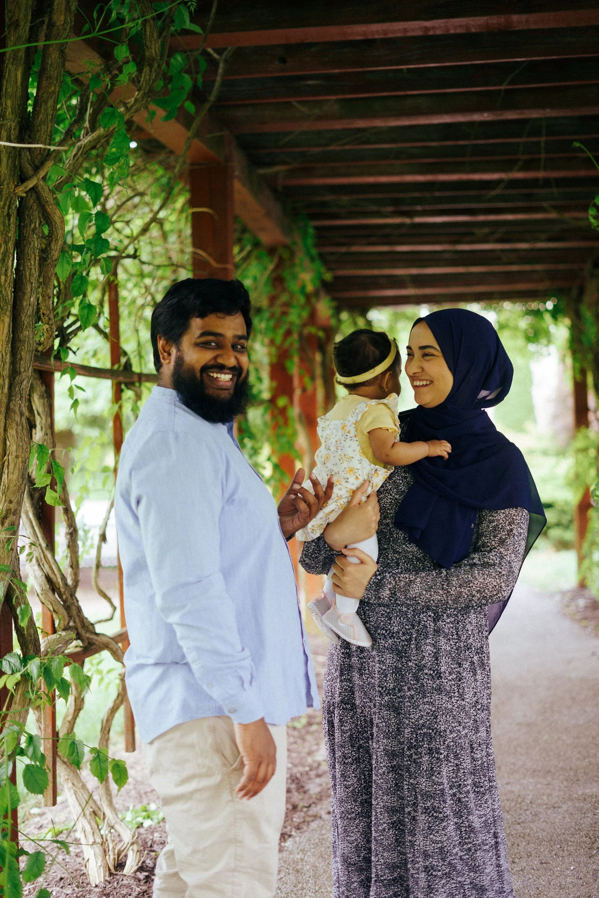 A smiling couple, a man with a beard and a woman wearing a dark blue hijab, are outdoors under a canopy of green vines. The woman is holding a baby girl dressed in yellow and white, and they are all enjoying a happy moment together.