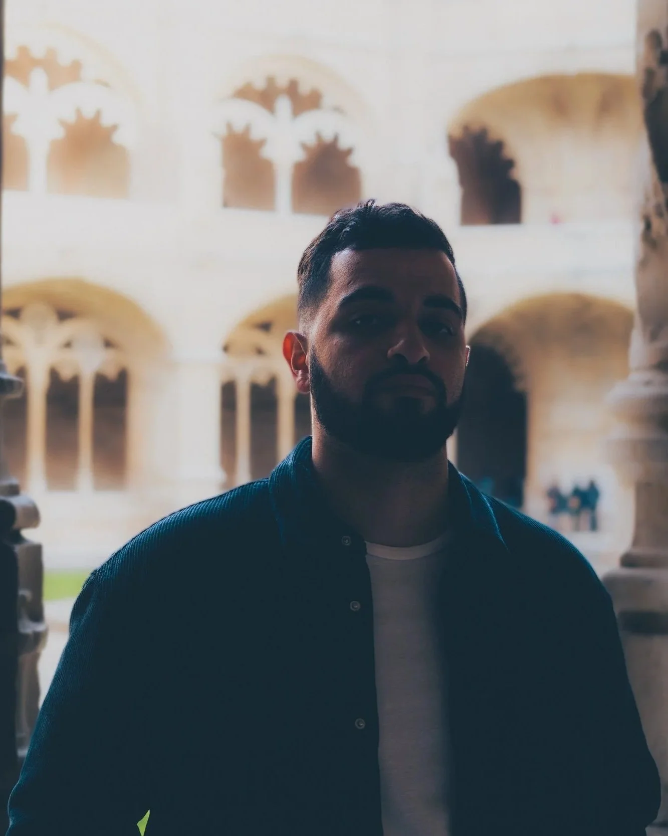 A man with a beard standing in front of a historic building with arched windows and intricate stonework.