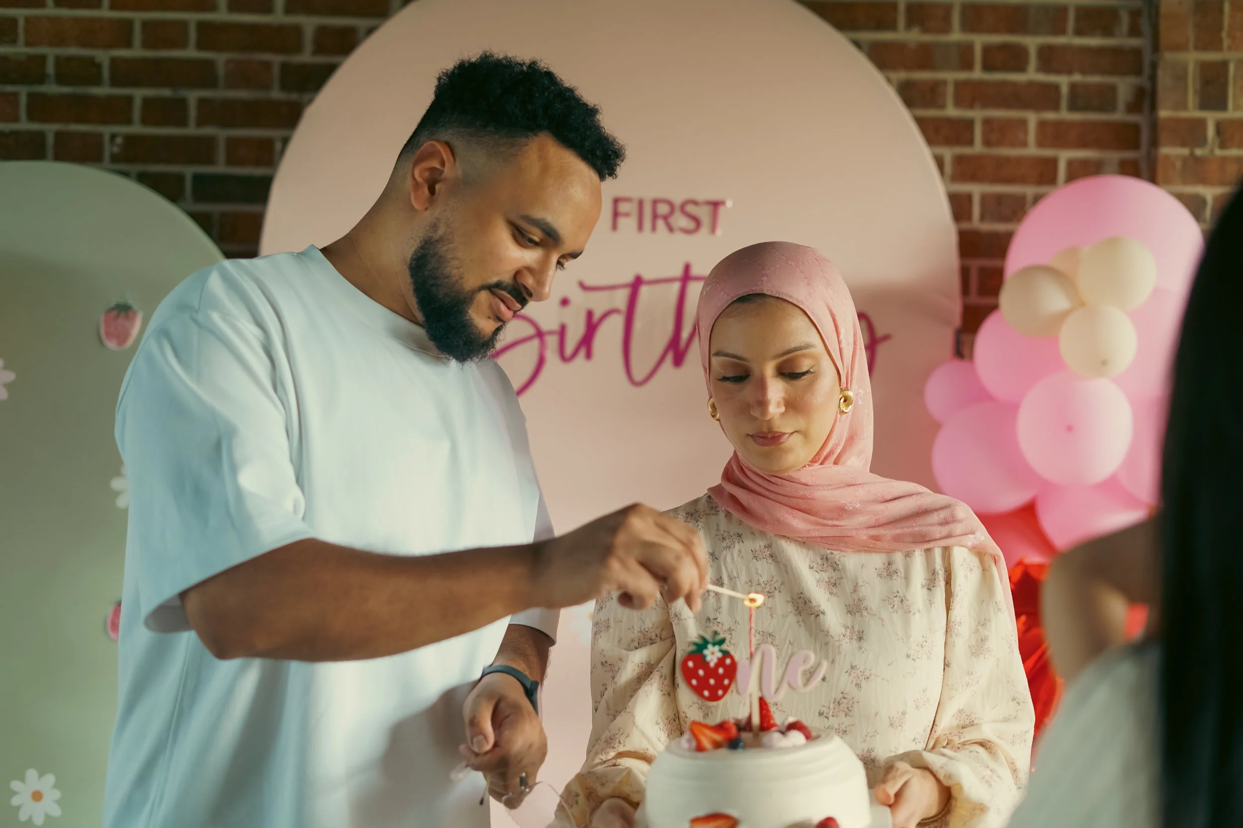 A man and a woman celebrating a birthday, with a cake decorated with strawberries and a strawberry-shaped cake topper, in front of a pink and white background with balloons.