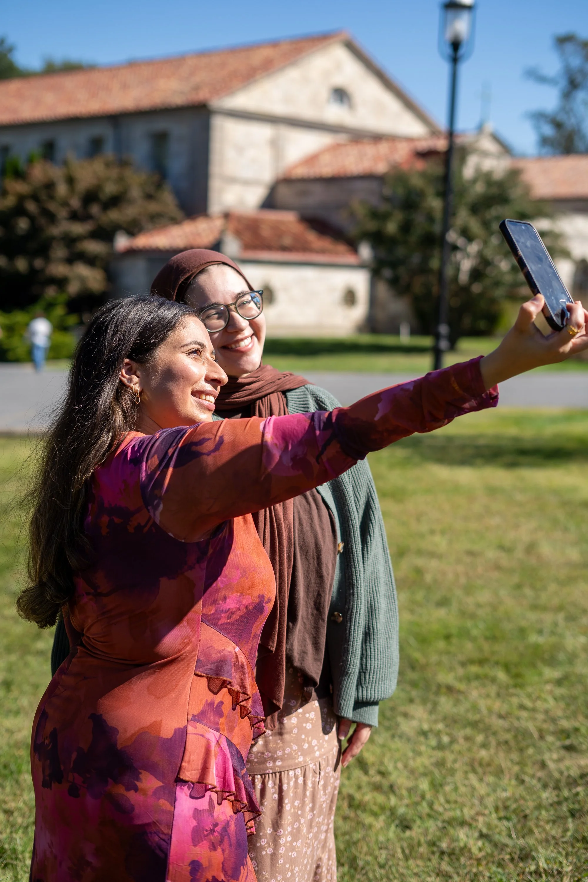 Two women taking a selfie outdoors on a sunny day, standing on grass with a historic building and trees in the background.