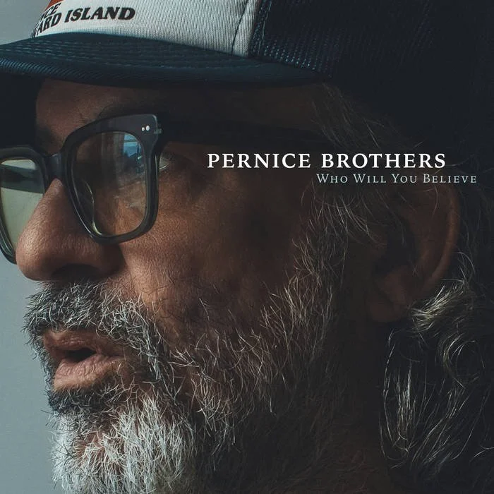 Close-up of a man wearing glasses and a cap, with a beard and mustache, looking to the left. Text on the image reads 'Pernice Brothers' and 'Who Will You Believe'.