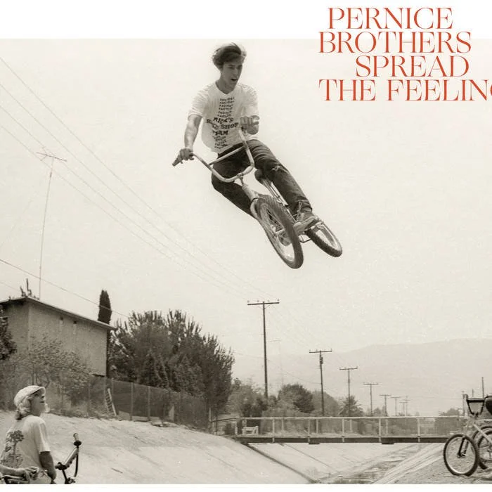 A black and white photo of a young man performing a aerial BMX trick in mid-air. He is wearing a t-shirt and jeans. A girl with a helmet is watching from below, and there are bike ramps and power lines in the background. In the top right corner, the text reads "Pernice Brothers Spread the Feeling."