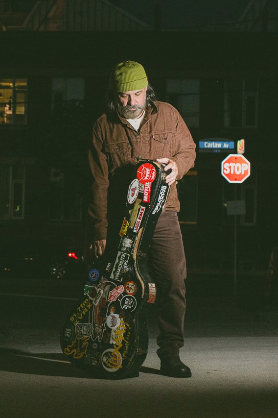 A man with long hair, glasses, and a beard standing at night on a street, wearing a green beanie and brown jacket, holding a guitar case covered with various stickers.