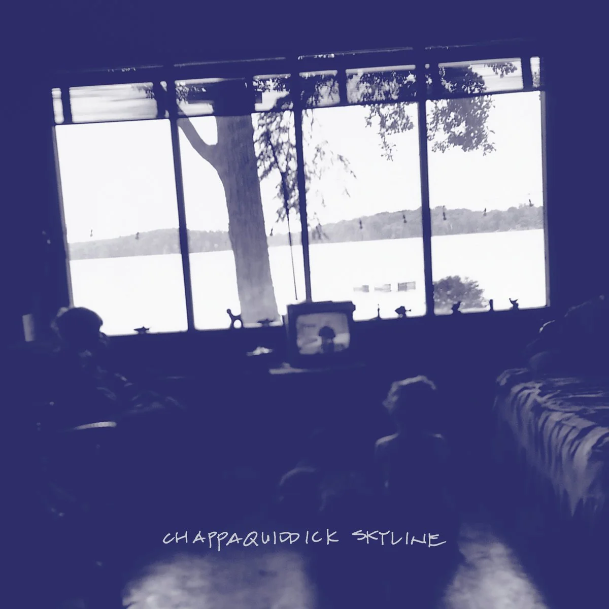 Interior view of a room with a large window overlooking a lake with trees and hills in the distance. Silhouettes of people are visible inside the room, and the scene is in black and white with the words "Chappaquiddick Skyline" at the bottom.