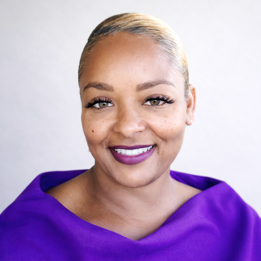 A smiling woman with short blonde hair, wearing a purple top, standing against a light background.