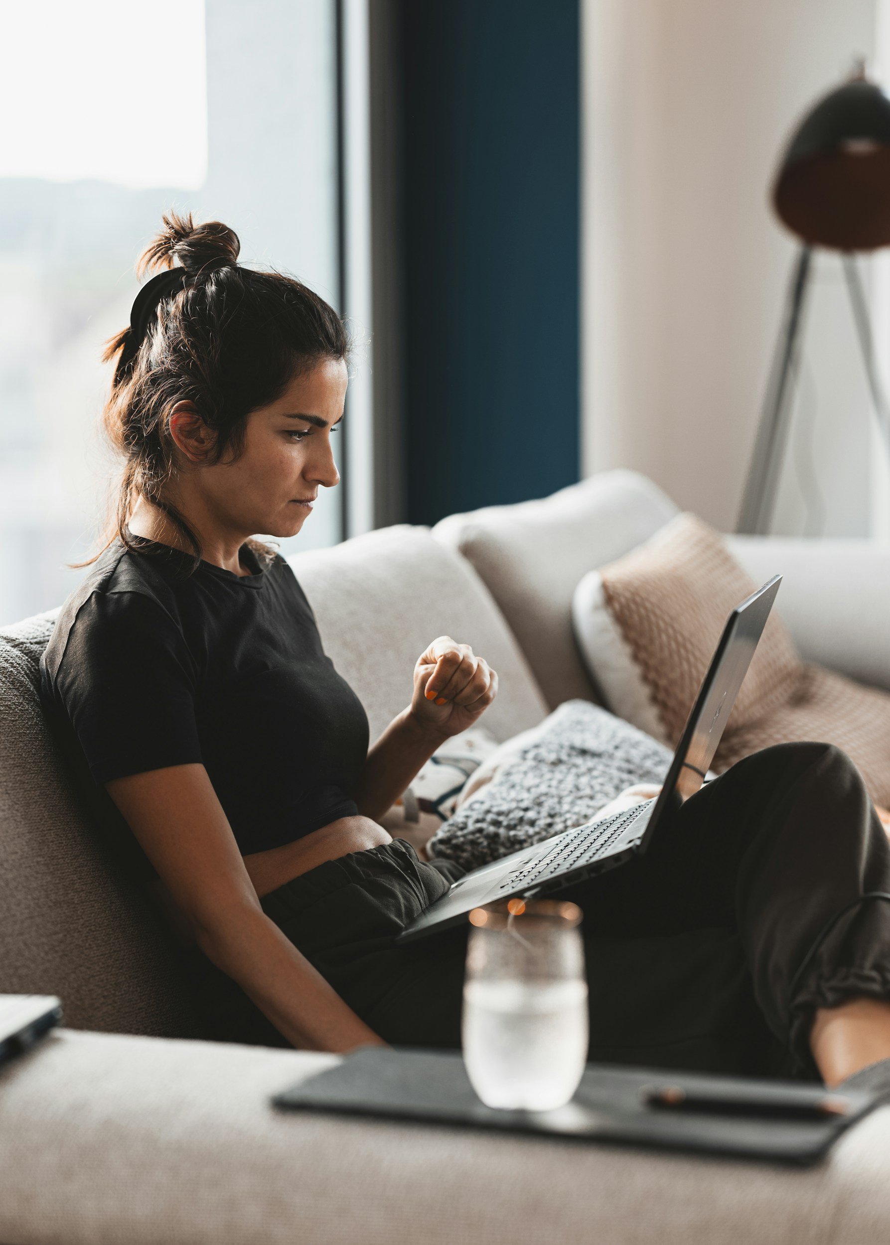 Young woman with dark hair tied in a bun, sitting on a beige couch, looking at a laptop with a determined expression, clenched fist, in a modern living room.