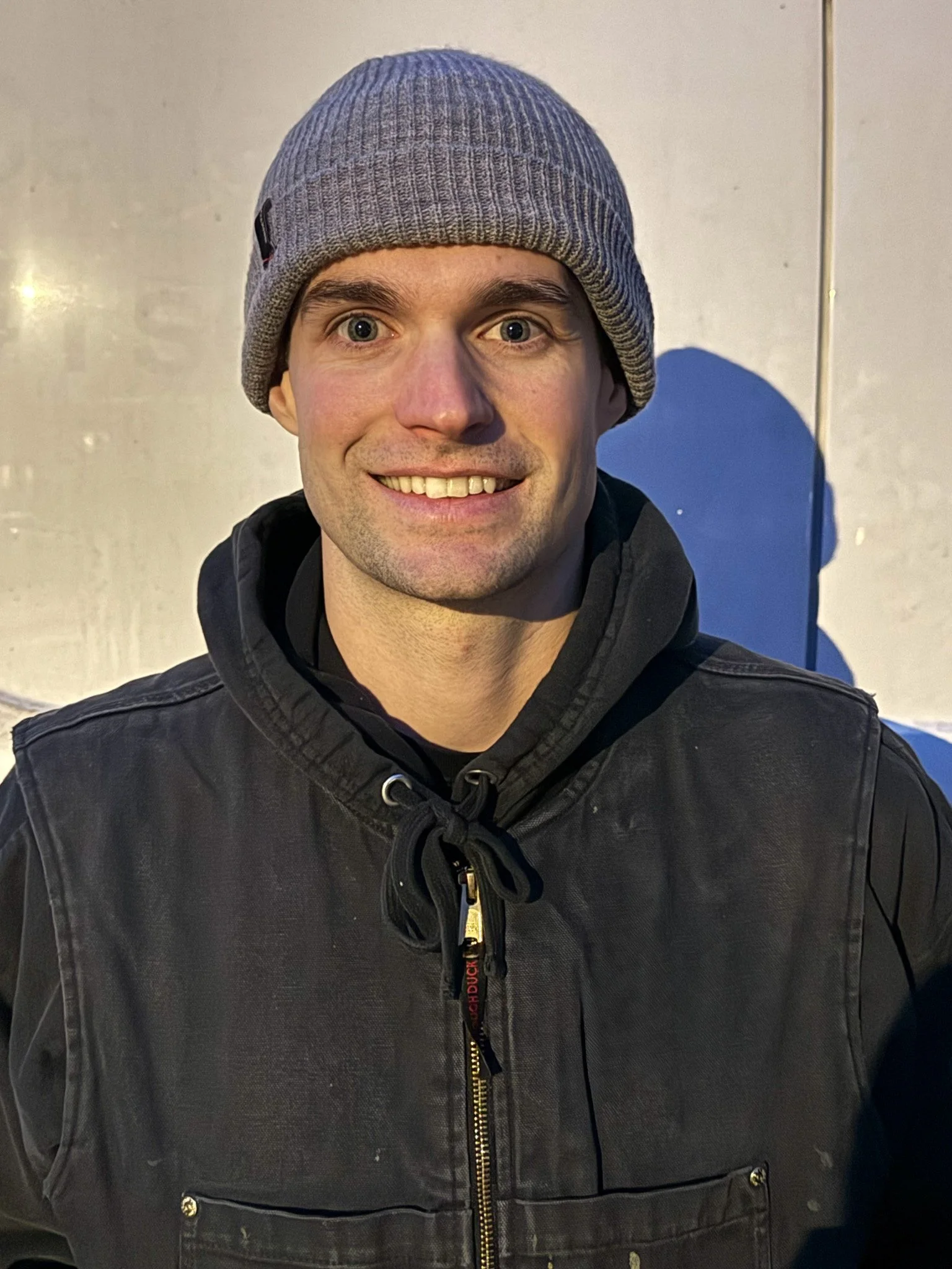 A smiling man wearing a gray beanie and a black jacket, standing against a plain wall with a shadow cast on it.