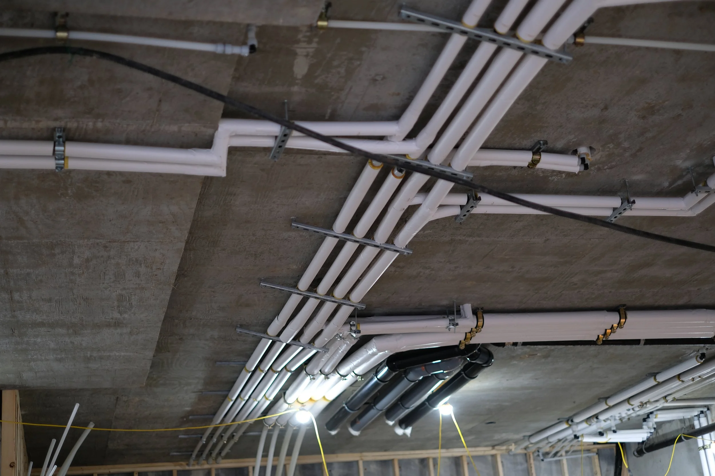 Indoor construction site with exposed concrete ceiling and white PVC piping for plumbing and electrical wiring, suspended by metal brackets, with yellow extension cords and hanging light bulbs.