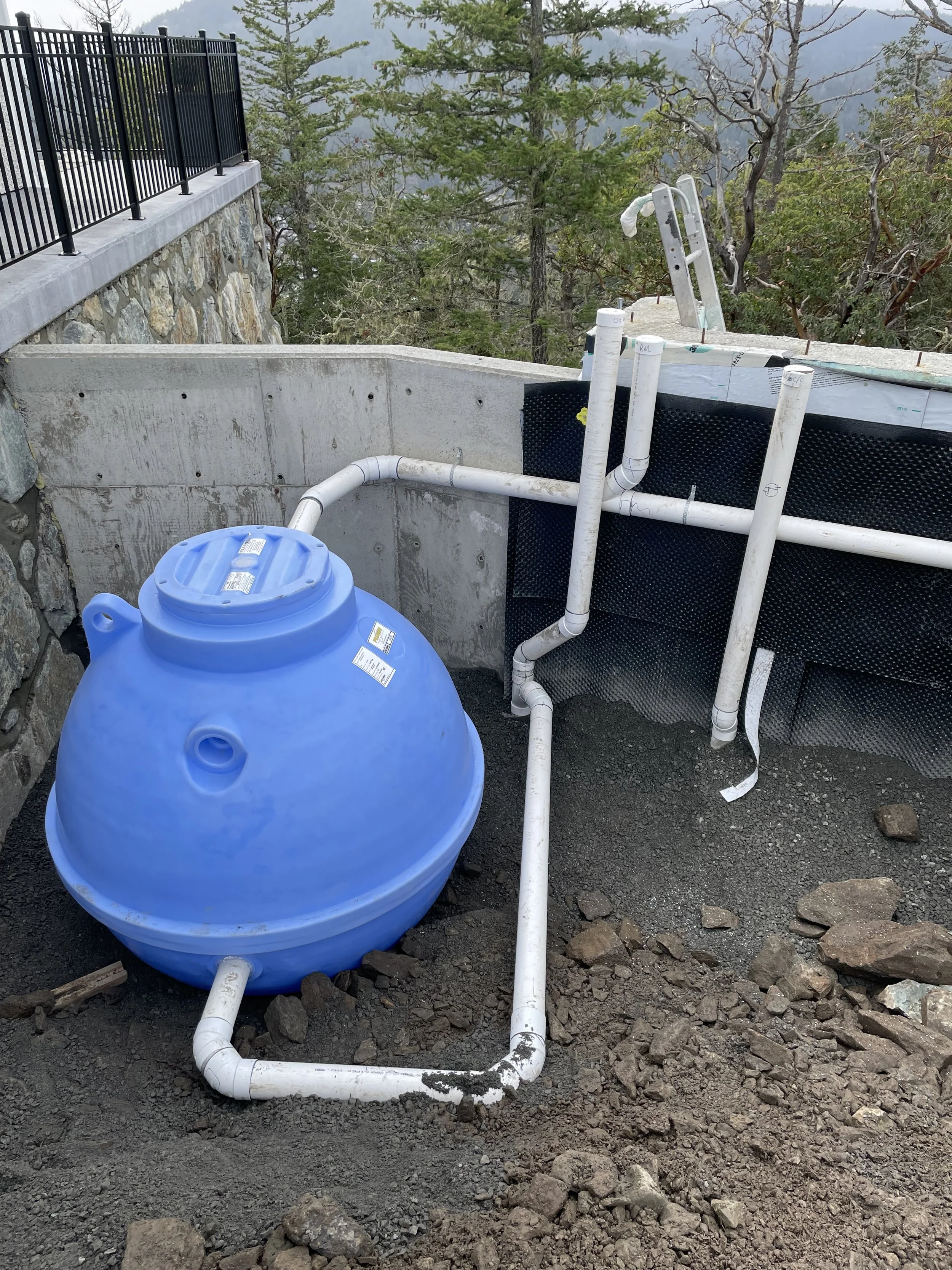 Construction site with a blue water tank, PVC pipes, and a concrete retaining wall, surrounded by trees and mountains in the background.