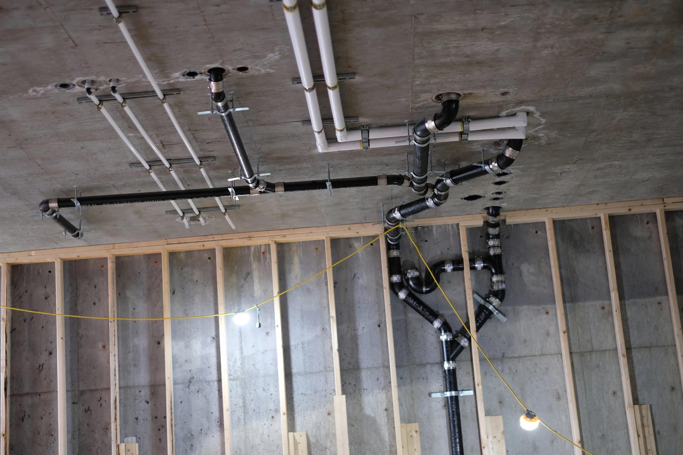 Construction site with exposed plumbing pipes attached to a concrete ceiling and wall, and wooden framing on the wall.