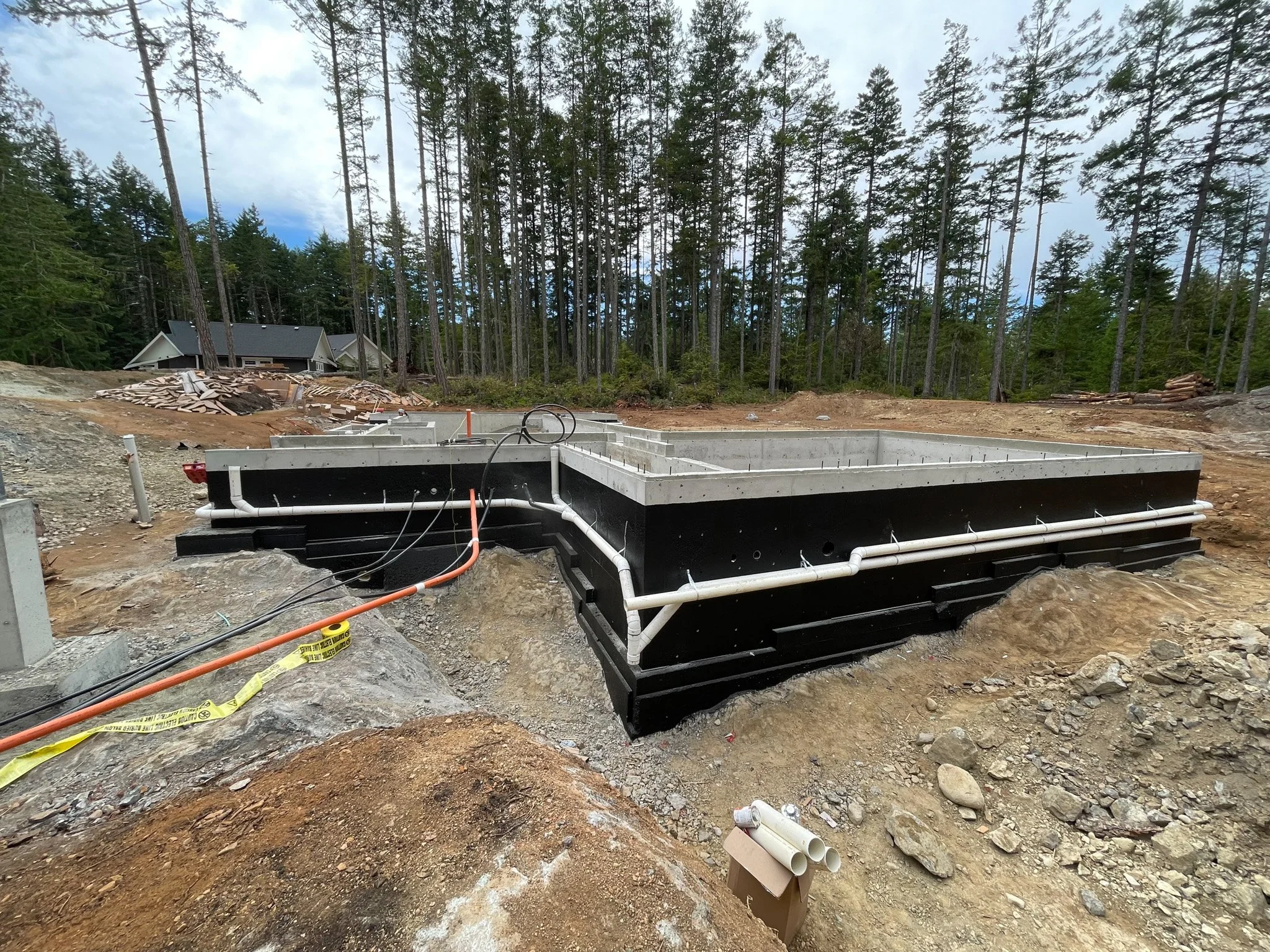 Construction site with a building foundation and pipes in a forested area, with a house visible in the background.