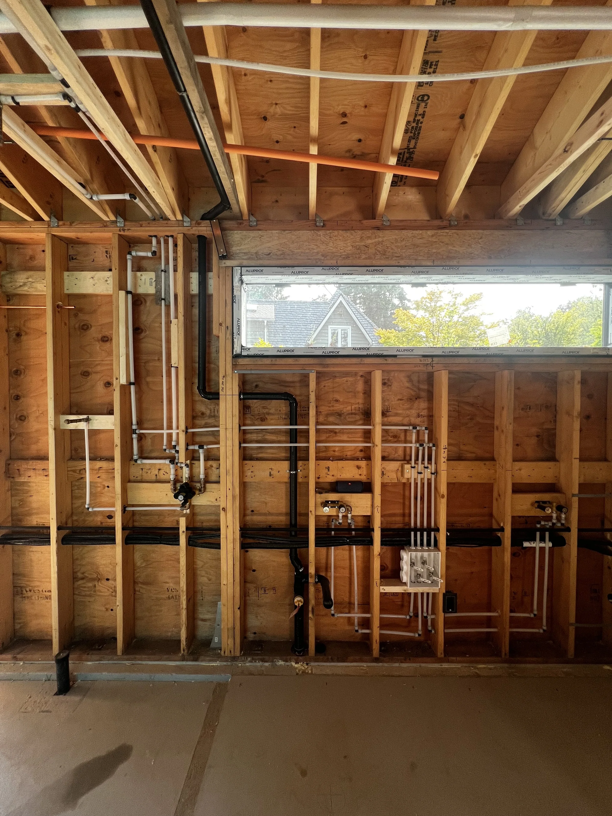 Interior construction of a building with exposed wooden framing and plumbing pipes, including black, white, and orange pipes, with a horizontal rectangular window showing trees and a house outside.