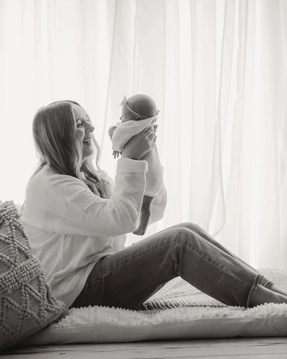 A woman sitting on a rug, holding a baby up in the air near a bright window with curtains, smiling and looking at the baby.