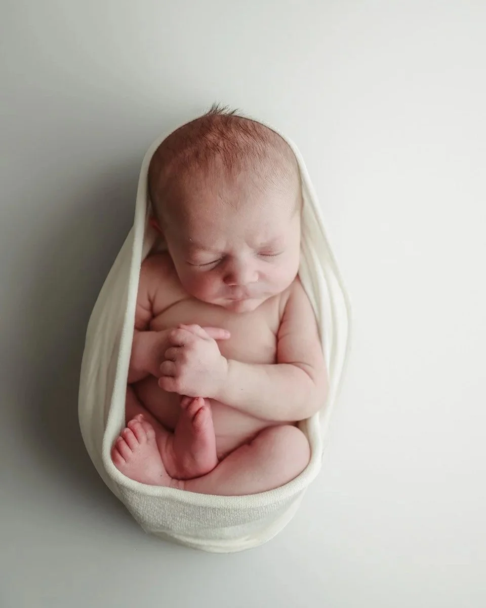 A sleeping newborn baby curled up inside a white cloth wrap, resting on a light-colored surface.