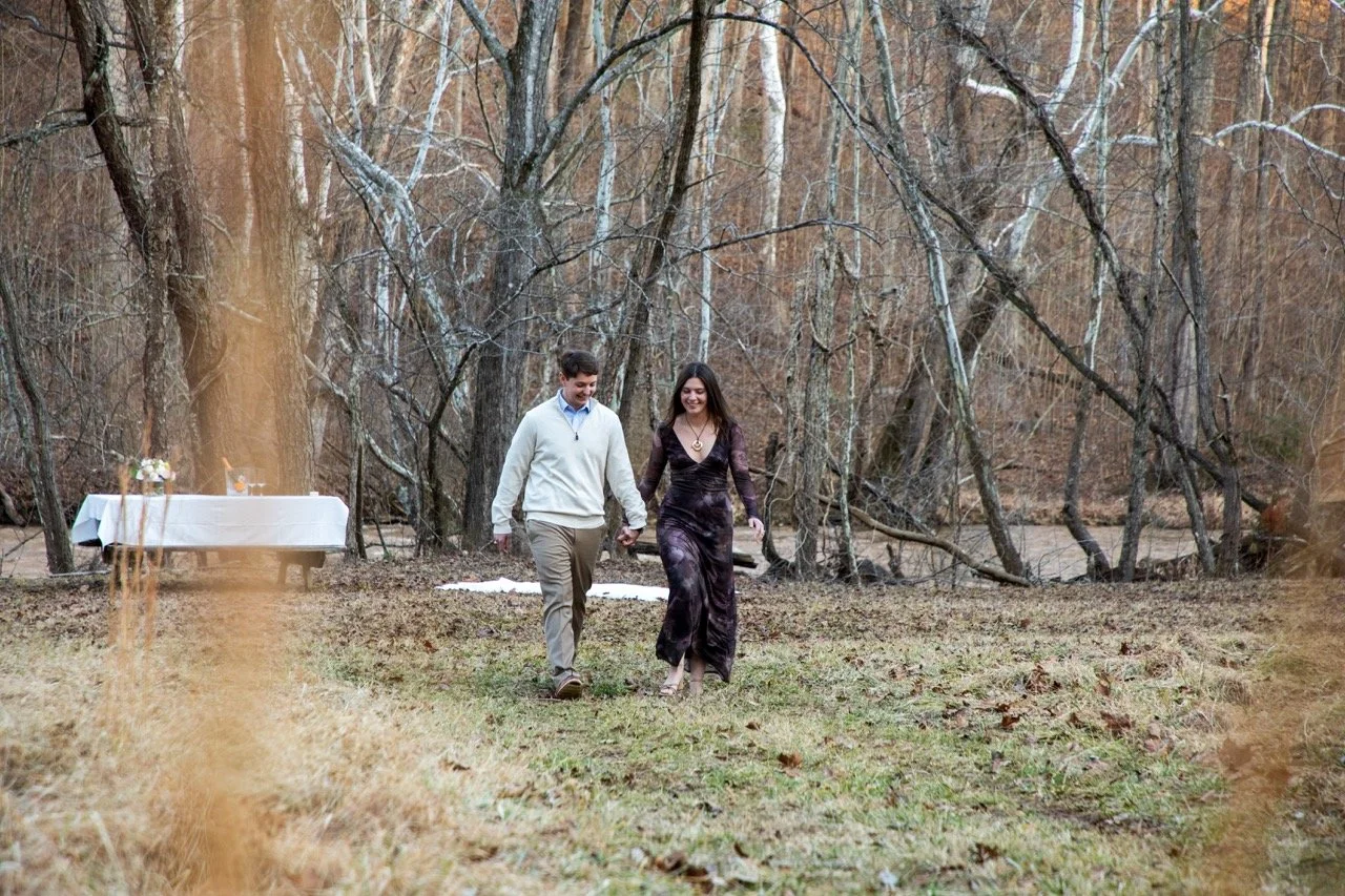 Caroline and Clay smiling as they walk hand-in-hand through a wooded field after an RVA surprise proposal.