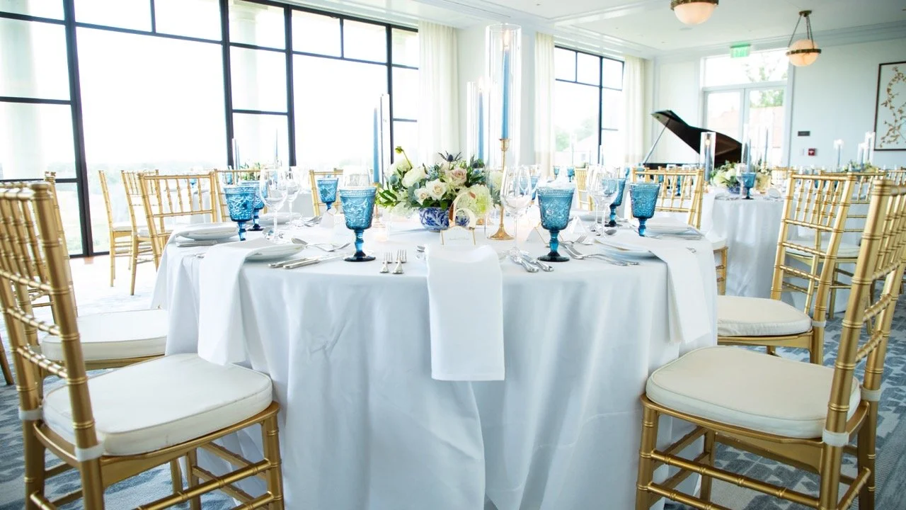 Bright, airy overhead view of white linen plates, modern utensils, and blue glassware at a custom dinner setup.