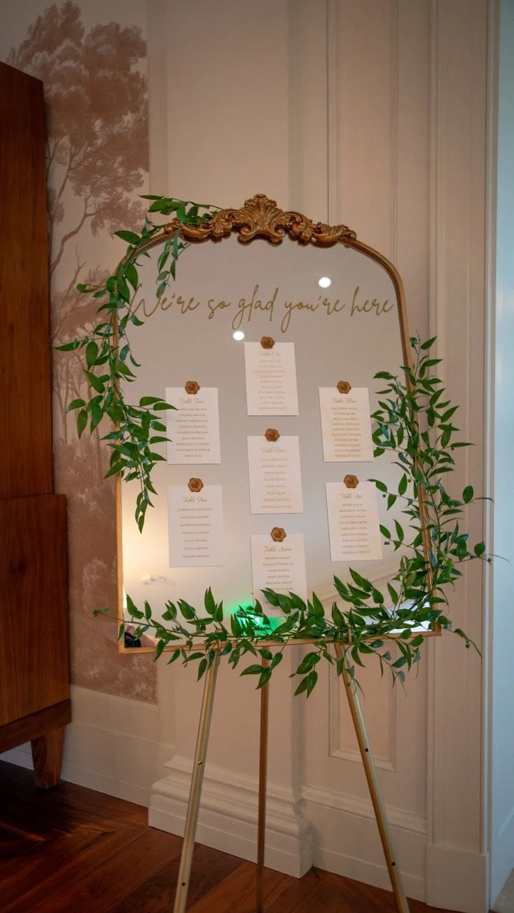 "We're so glad you're here" gold script mirror seating chart framed by creeping green vines at a wedding venue entrance.