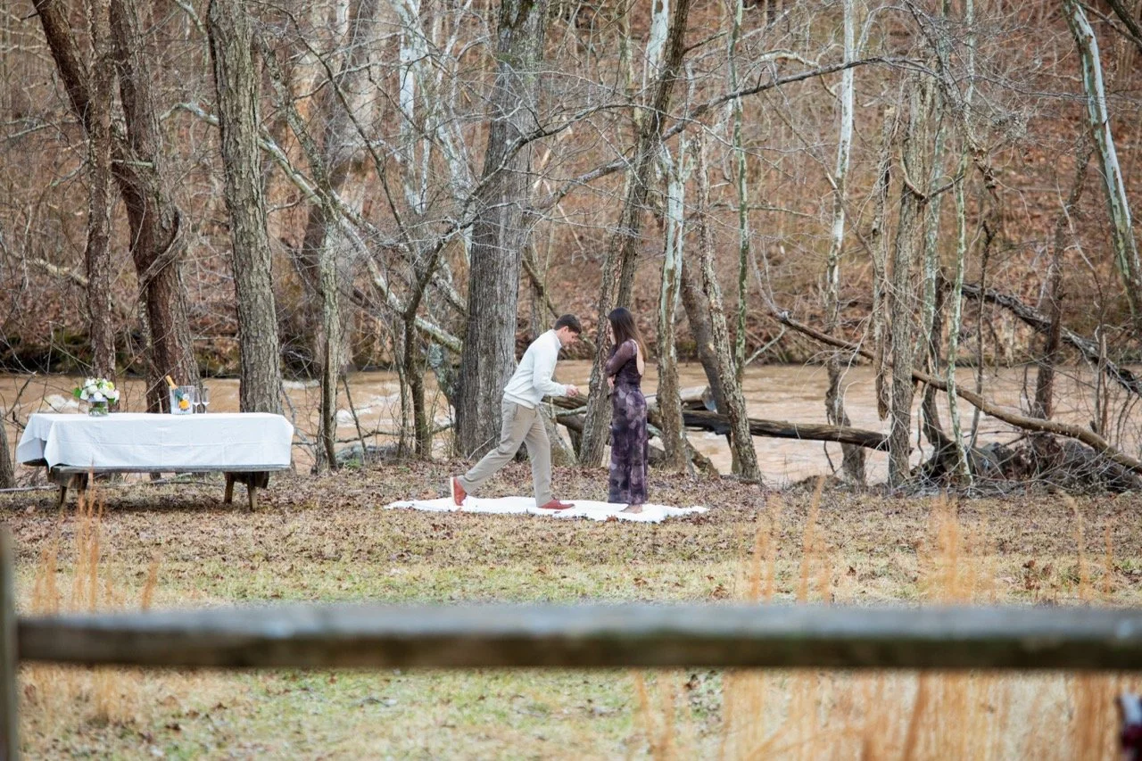 Clay getting down on one knee to propose to Caroline on a white blanket in the woods, captured by a Richmond proposal photographer.