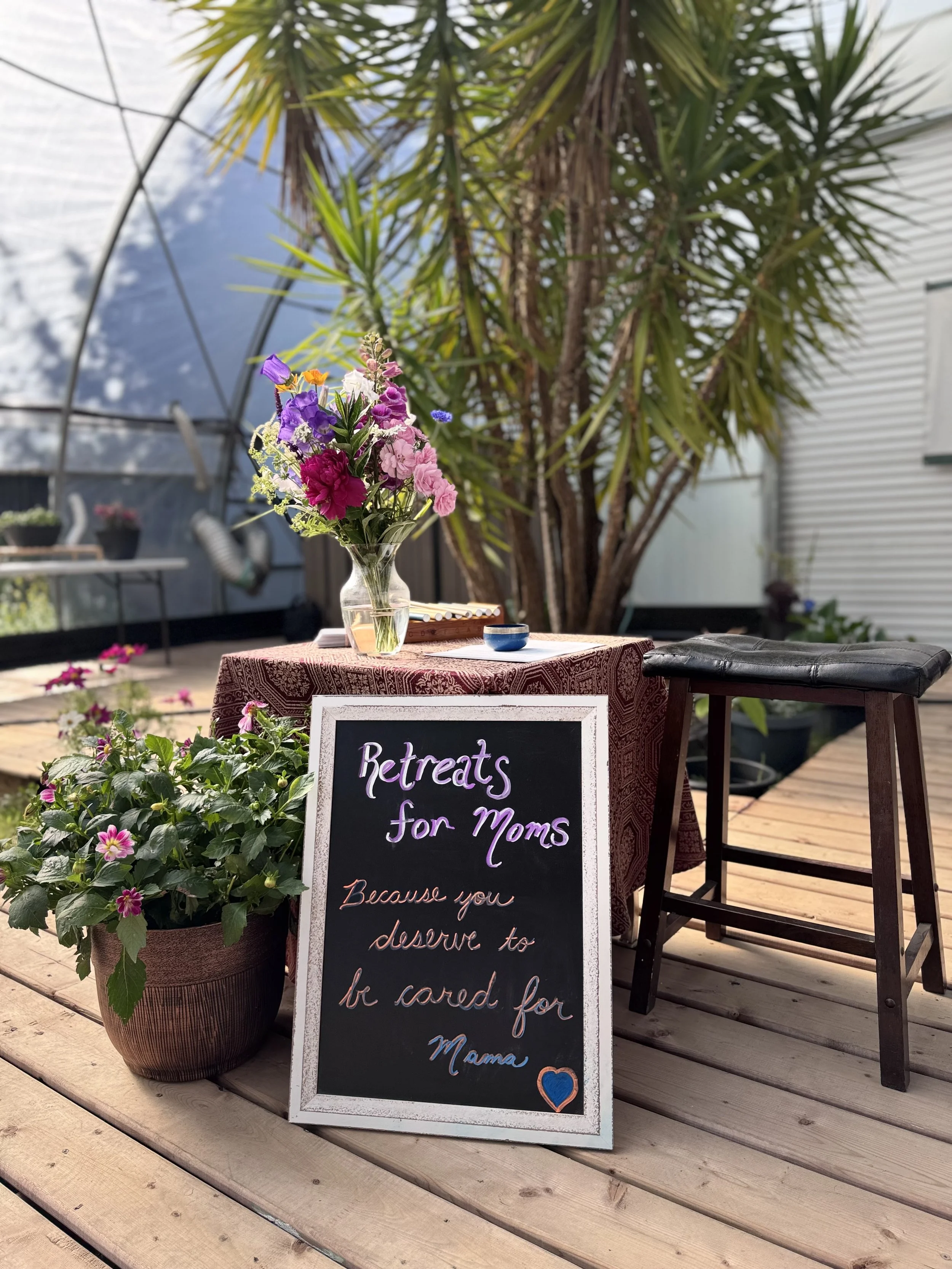 A welcoming setup for a mother’s retreat, featuring a table with a bouquet of colorful flowers, a chalkboard sign with a heartfelt message, and lush greenery in the background.