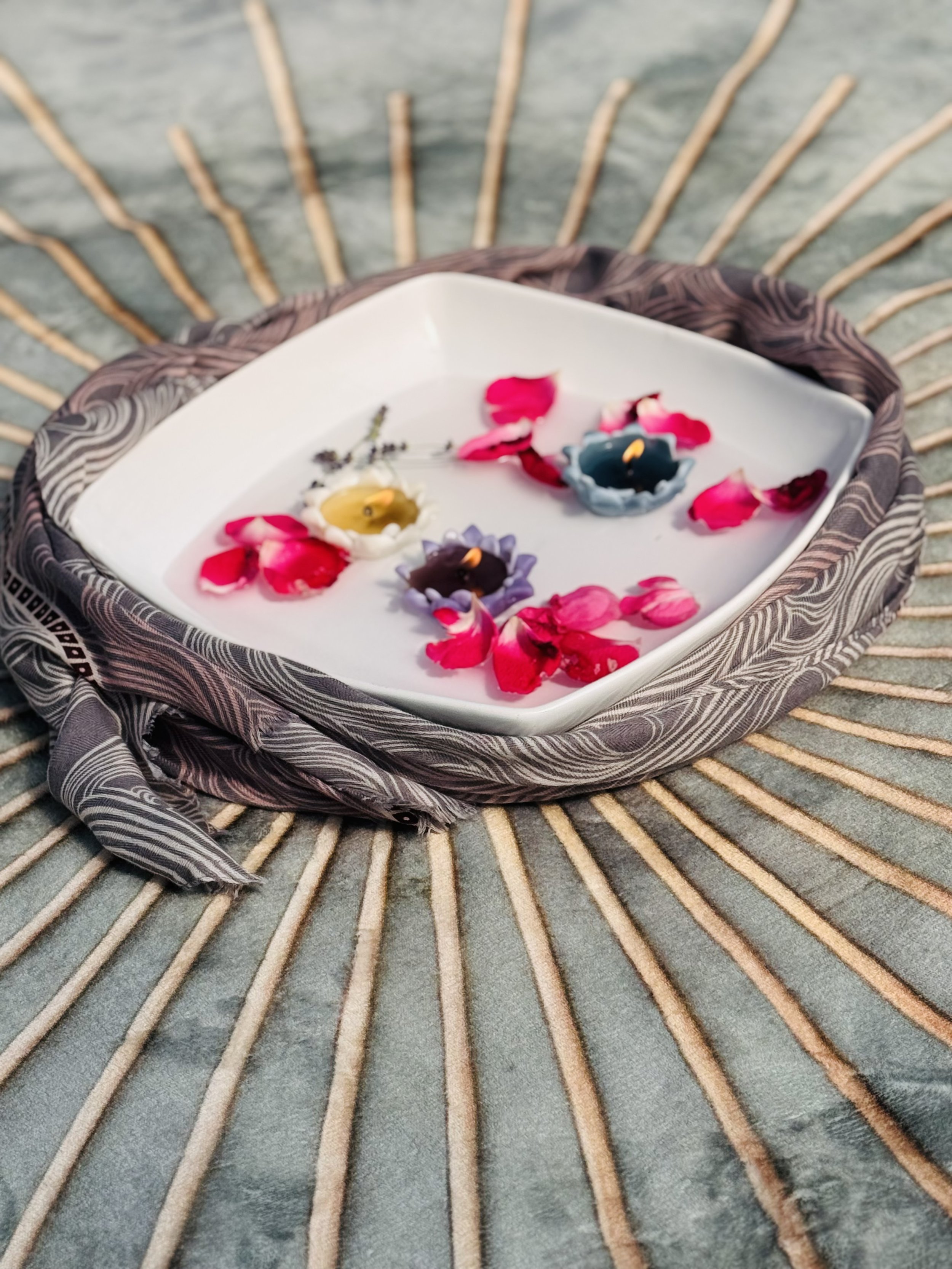 Decorative candles placed on a white dish with rose petals inside, all resting on a fabric with a striped pattern, in front of a background with radiating wooden sticks.