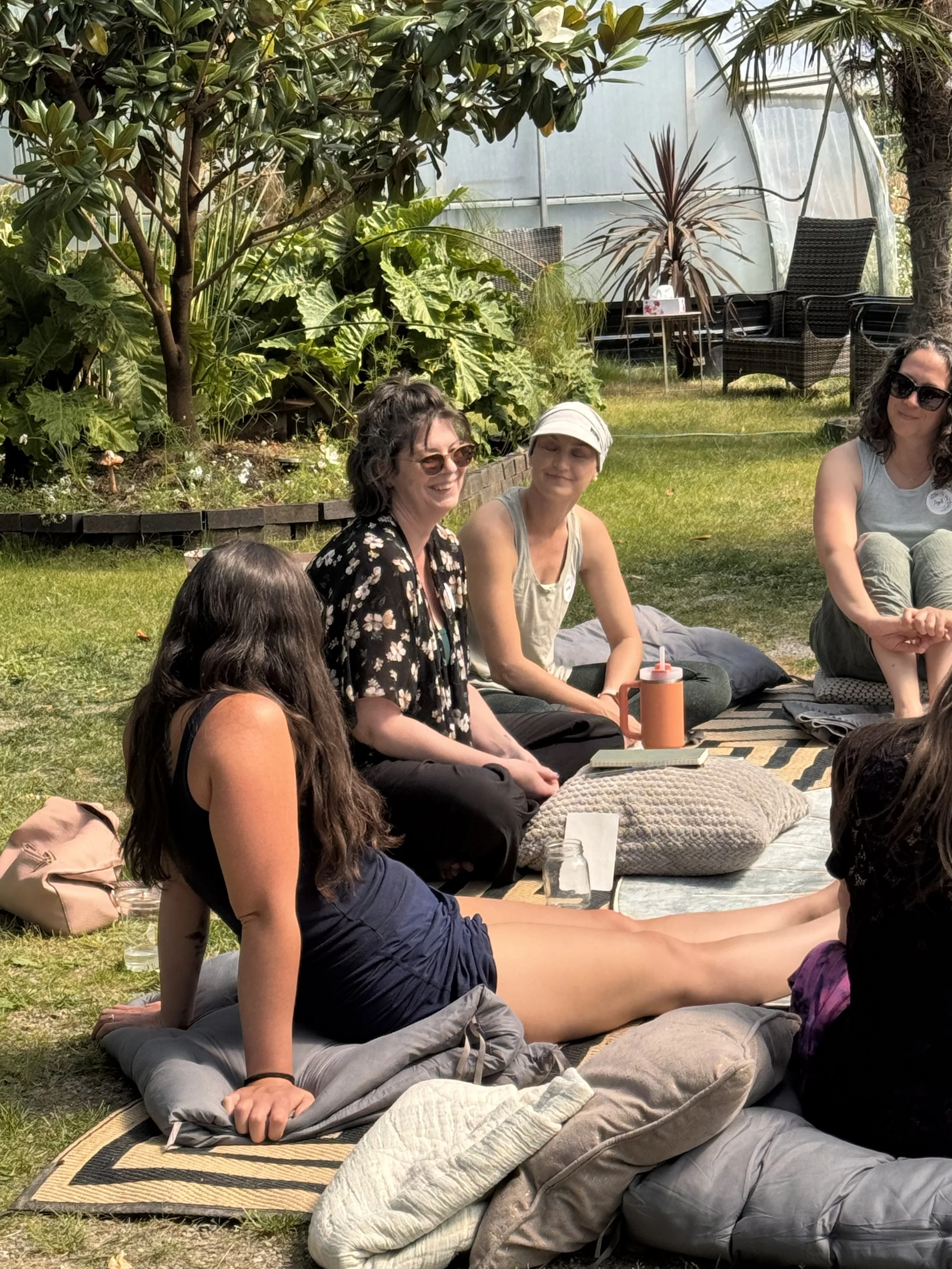 Group of women sitting outdoors on a blanket, engaged in conversation, surrounded by greenery and plants in a garden setting.