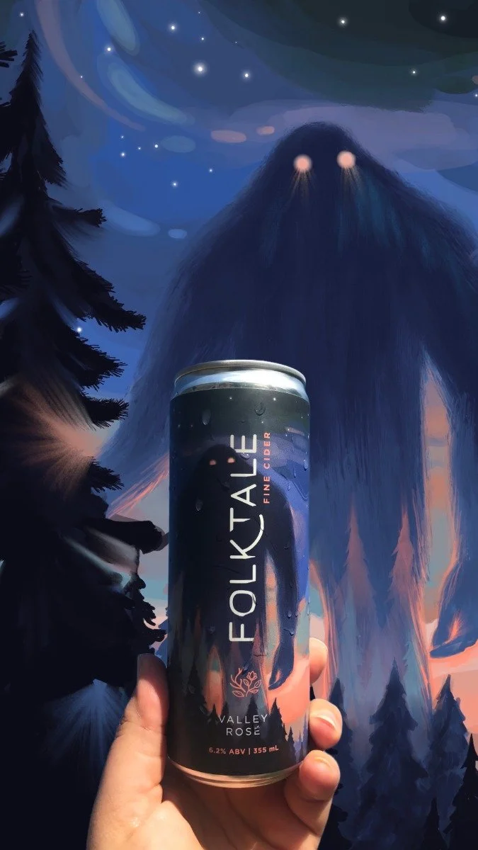 A hand holding a can of Folk Tale Valley Rosé cider in front of a digital or painted background depicting a mountain with glowing eyes and a starry night sky.