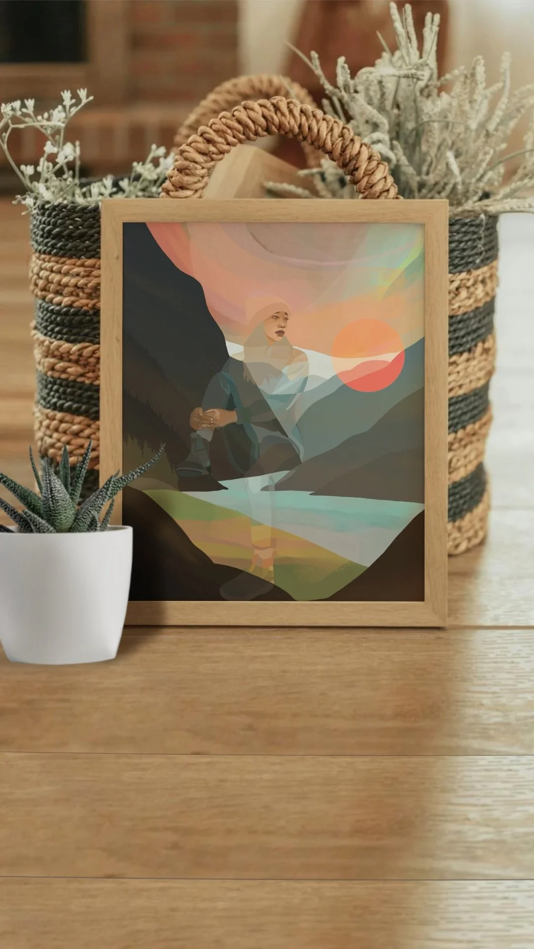 Digital art of a woman with a hijab, sitting in a mountainous landscape with a sunset, displayed in a wooden frame in front of a plant and a woven basket.