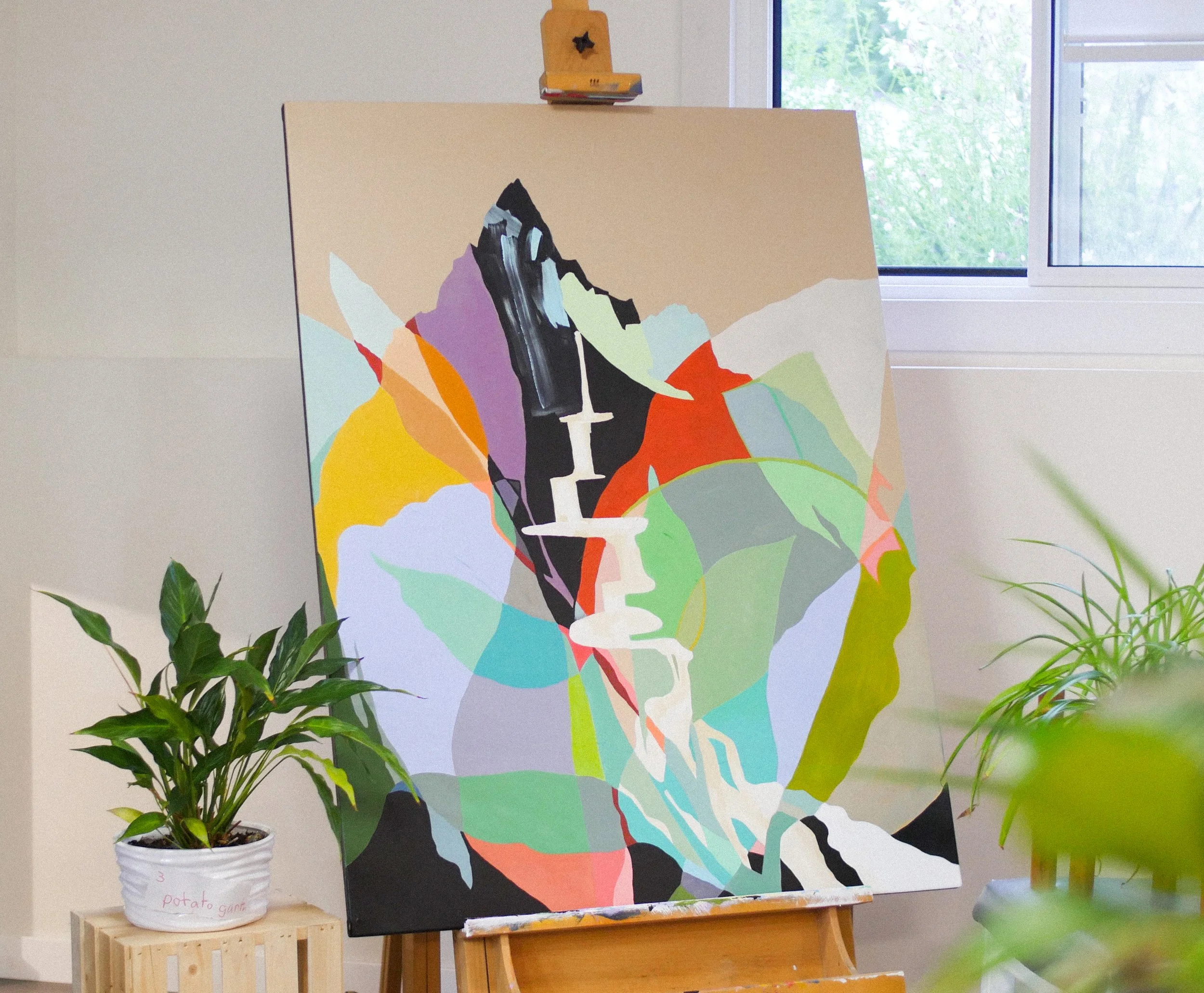 Colorful abstract painting on an easel next to potted plants and a window in a bright room.