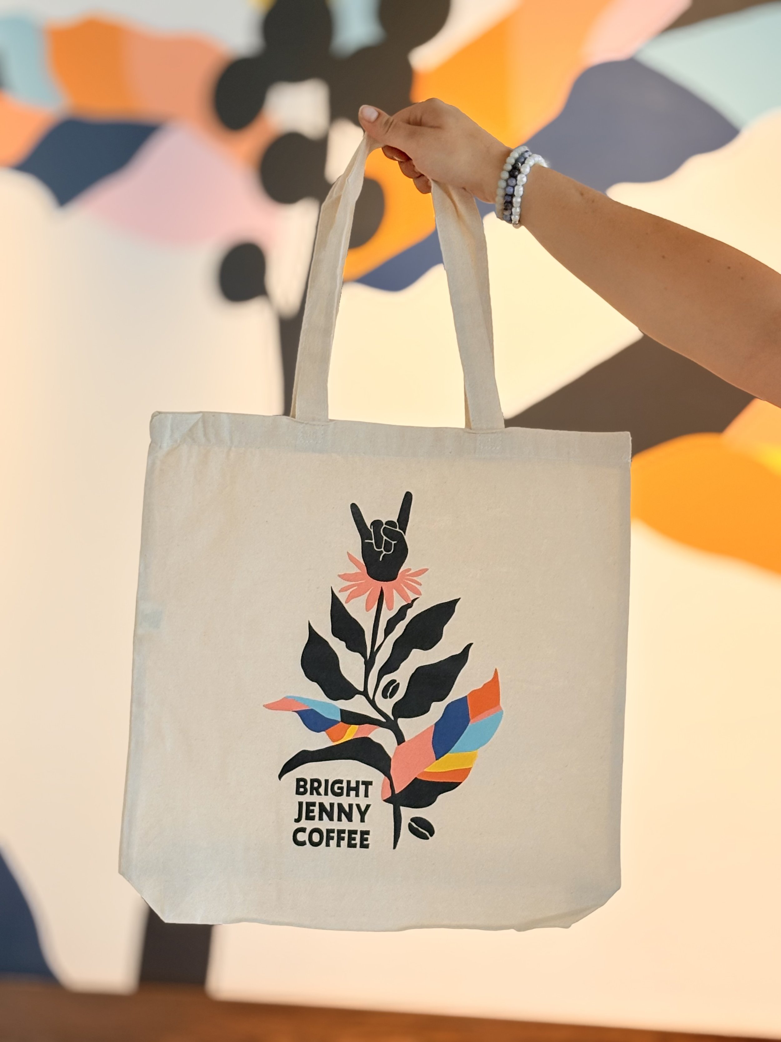 A white tote bag with a colorful floral design and the words "Bright Jenny Coffee" on it, being held up by a person with a beaded bracelet, against a background with abstract, colorful shapes.