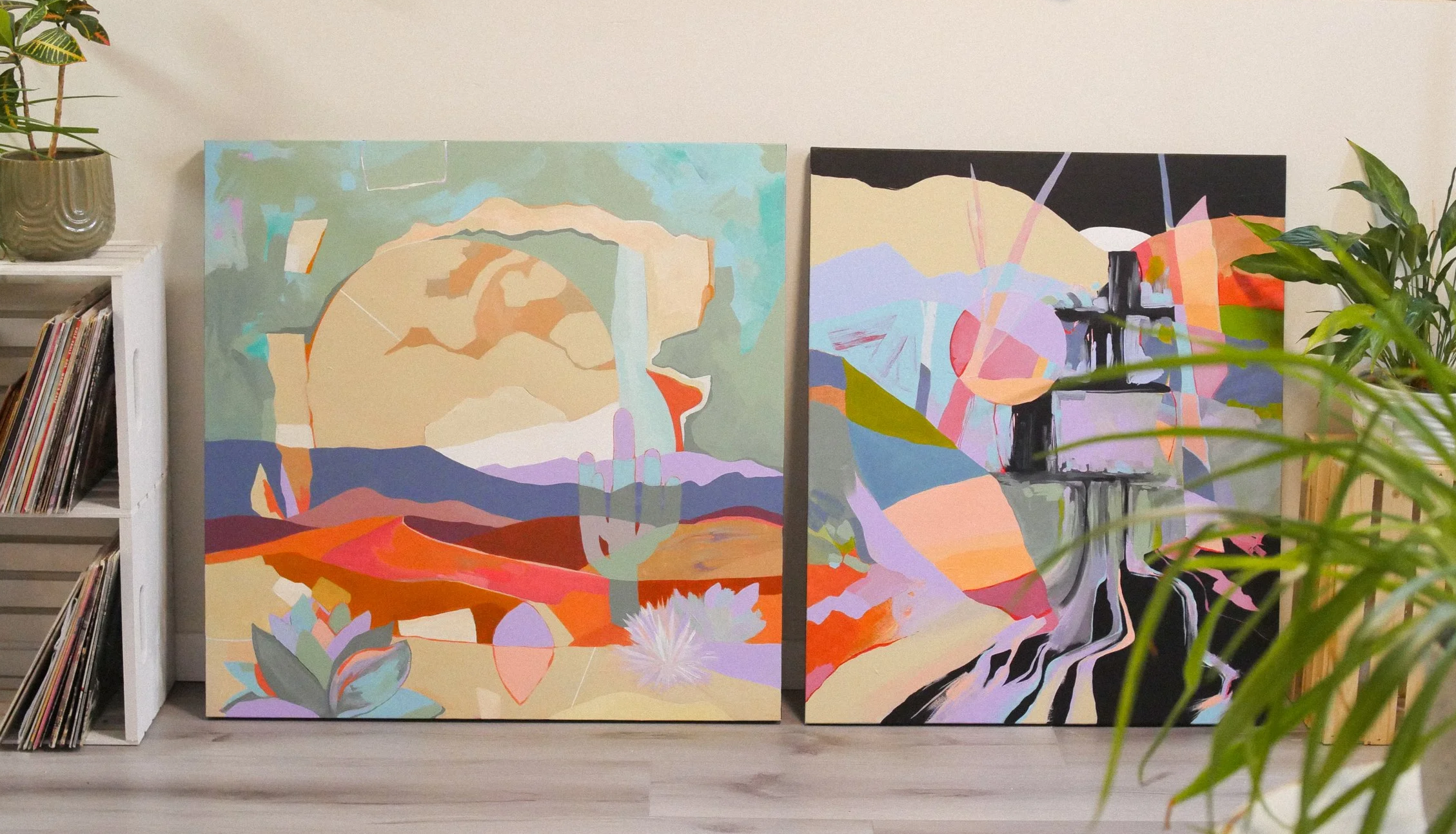 Two colorful abstract landscape paintings leaning against a white wall, with a potted plant on top of a bookshelf on the left and a green leafy plant on the right.