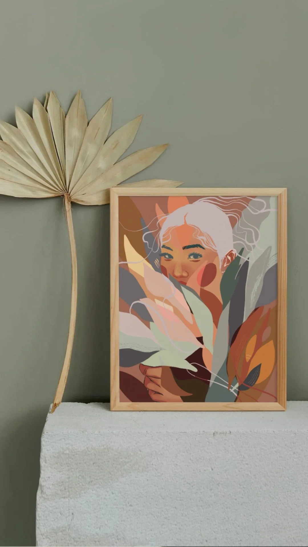 Framed abstract portrait of a woman with white hair, surrounded by colorful leaves and floral elements, placed on a white textured surface against a green wall, with a dried palm leaf on the left.