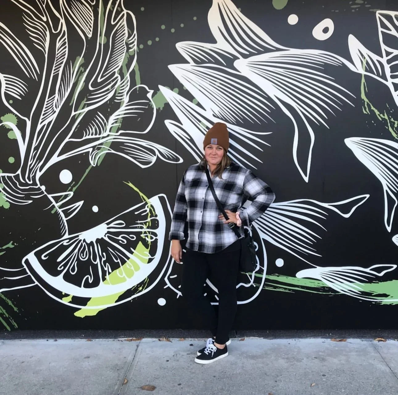 A woman wearing a brown beanie, black and white plaid shirt, black pants, and black sneakers with white soles, standing in front of a black mural with white and green line art of leaves and a lemon slice.