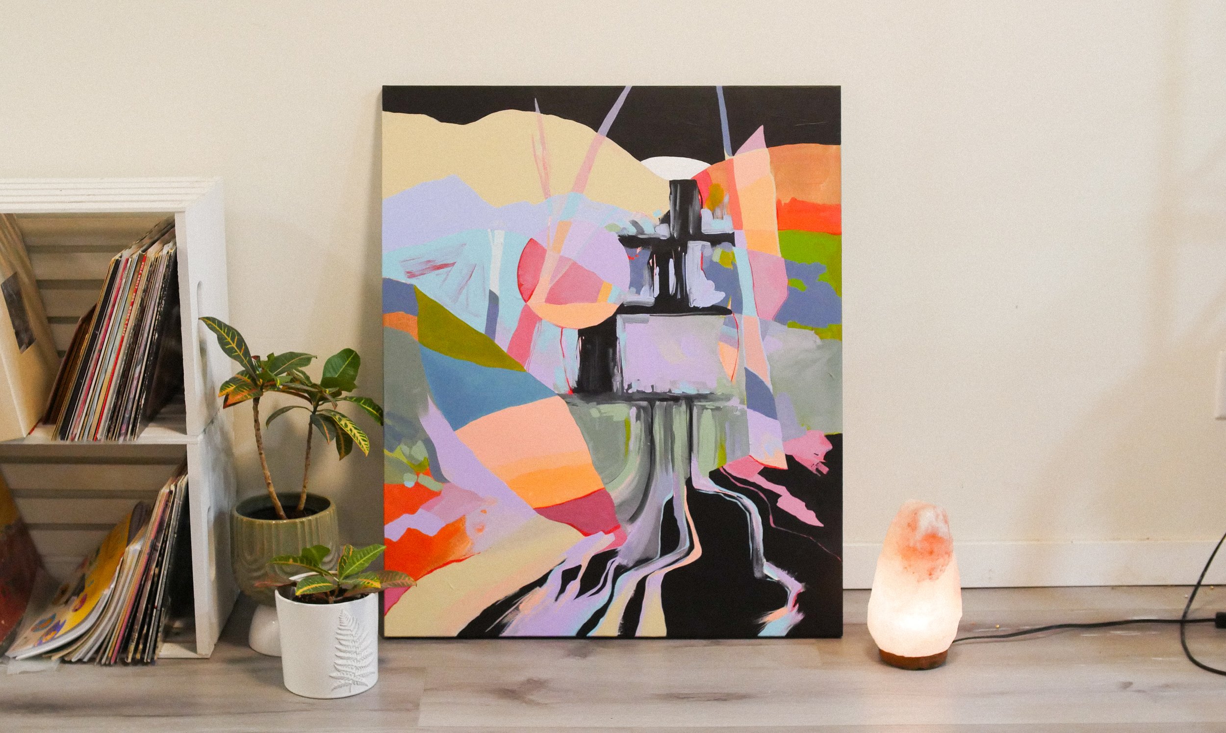 Abstract modern painting with bright colors and a cross in the center, leaning against a white wall. To the left are two potted plants of different sizes, one in a beige pot and one in a white pot. To the right, a salt lamp with an orange glow plugge
