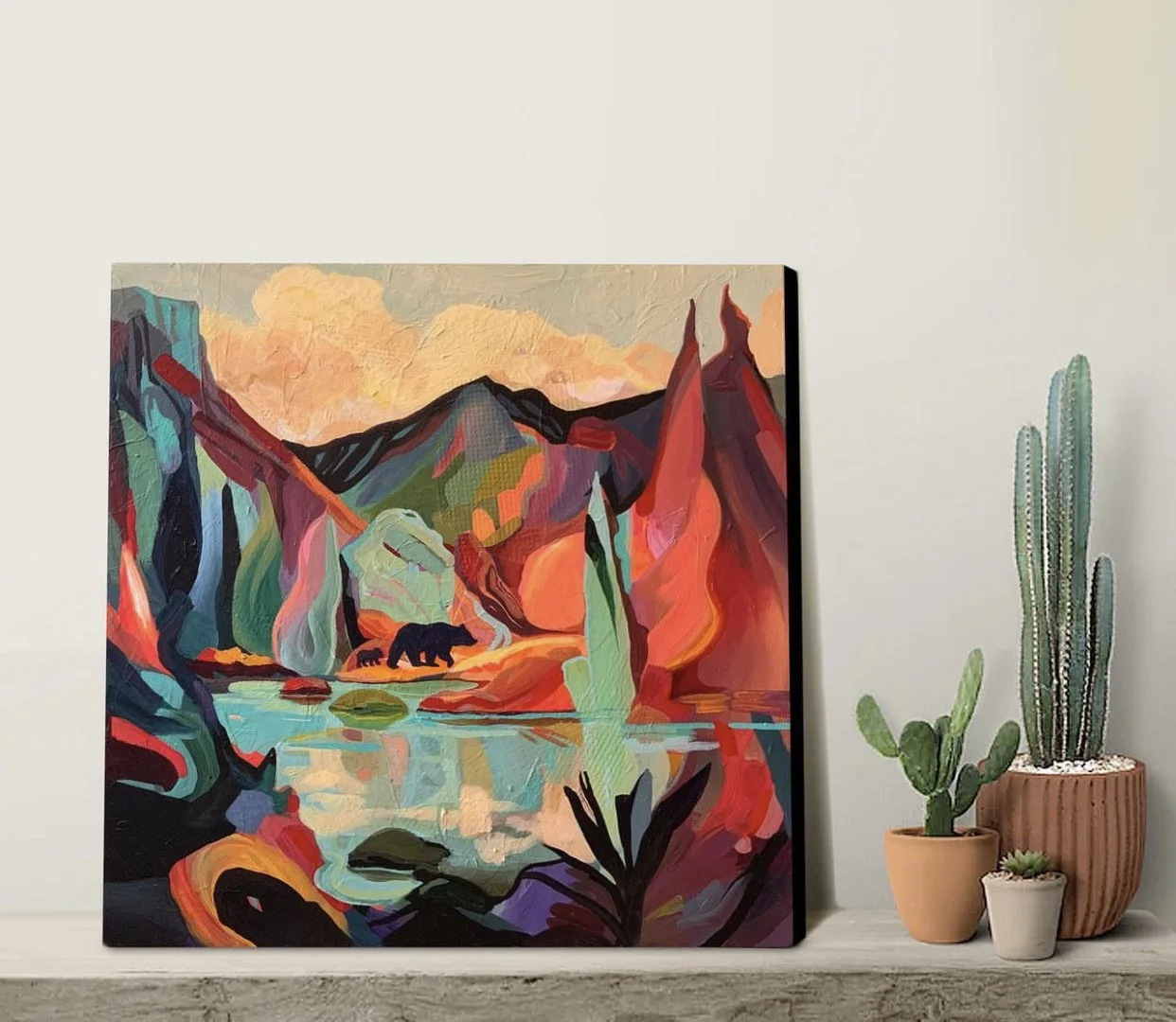 Colorful abstract landscape painting with mountains, a river, and a bear, placed on a shelf next to potted cacti and succulents.