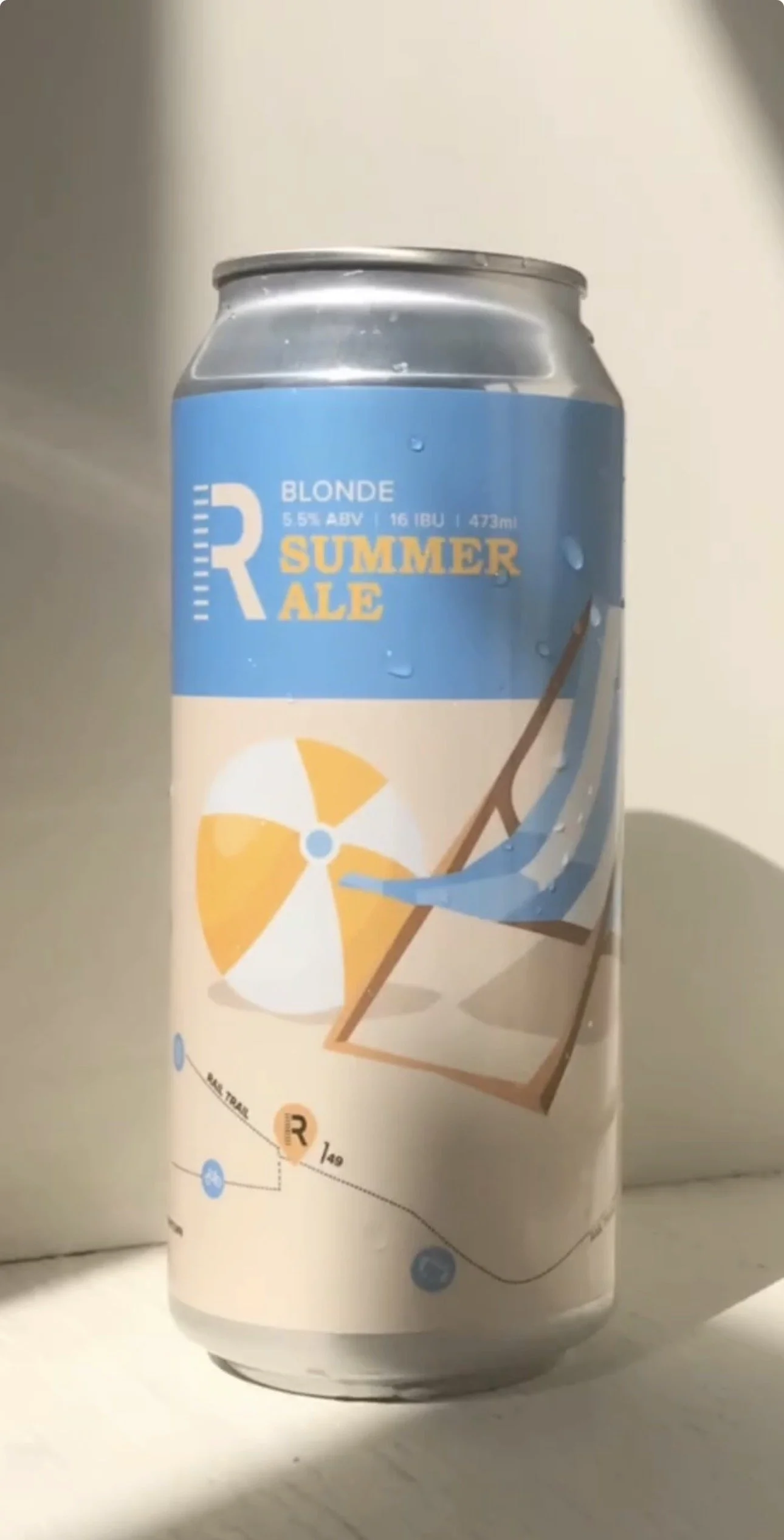 Can of Blonde Summer Ale with a blue and beige label, featuring a beach chair, umbrella, and surfboard, indicating a seasonal summer beer.